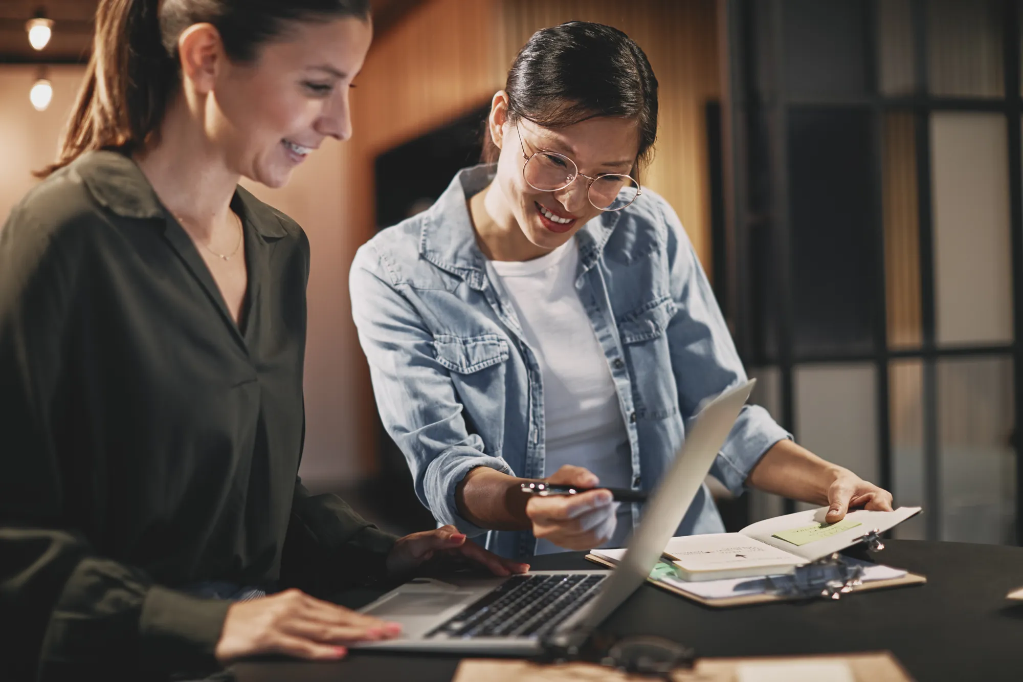 Two people smiling over a laptop and notebook in a warmly lit office